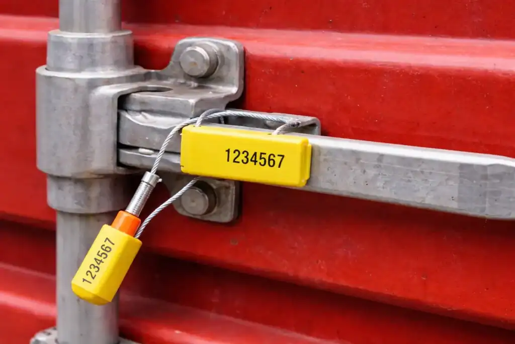 סגר מכולה (Container Seal) צהוב עם מספר סידורי המחובר לדלת מכולת מטען אדומה, משמש לאבטחת המכולה בזמן שינוע לוגיסטי.